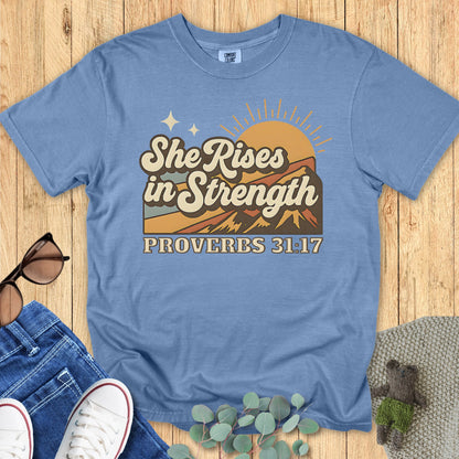 Flatlay of a Comfort Colors 1717 blue jean “She Rises in Strength – Proverbs 31:17” t-shirt with warm-toned mountain sunrise design. A faith-filled women’s scripture tee — durable faith-based apparel for women and encouraging modern Christian shirt, great for Christian mom gift ideas.