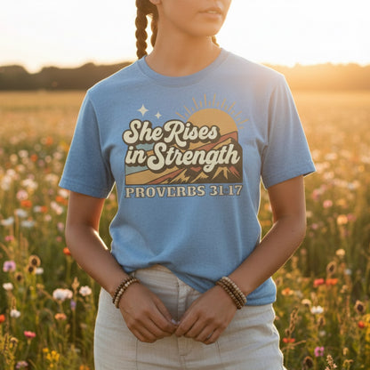 Woman standing in a sunlit field wearing a Comfort Colors 1717 blue jean “She Rises in Strength – Proverbs 31:17” t-shirt with a retro sunrise mountain design. A bold women’s scripture tee — faith-based apparel for women and empowering modern Christian shirt, perfect Christian mom gift idea.