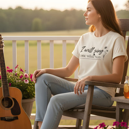 Woman sitting on a porch at sunset wearing a Comfort Colors 1717 ivory “I Will Sing Come What May” t-shirt with delicate floral and bird line art. A peaceful women’s scripture tee — uplifting faith-based apparel for women and inspiring modern Christian shirt, ideal Christian mom gift idea.