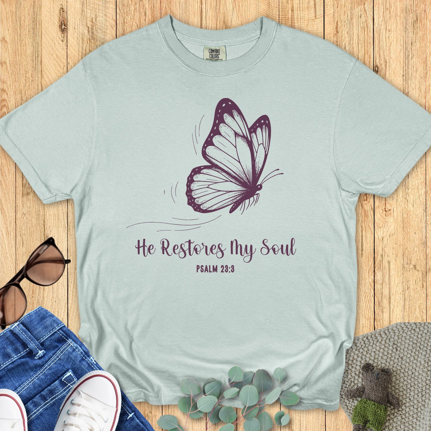 Flatlay of a Comfort Colors 1717 bay “He Restores My Soul – Psalm 23:3” t-shirt in soft mint tone with graceful butterfly print. A calming women’s scripture tee — modern faith-based apparel for women and meaningful Christian mom gift idea inspired by Psalm 23:3.