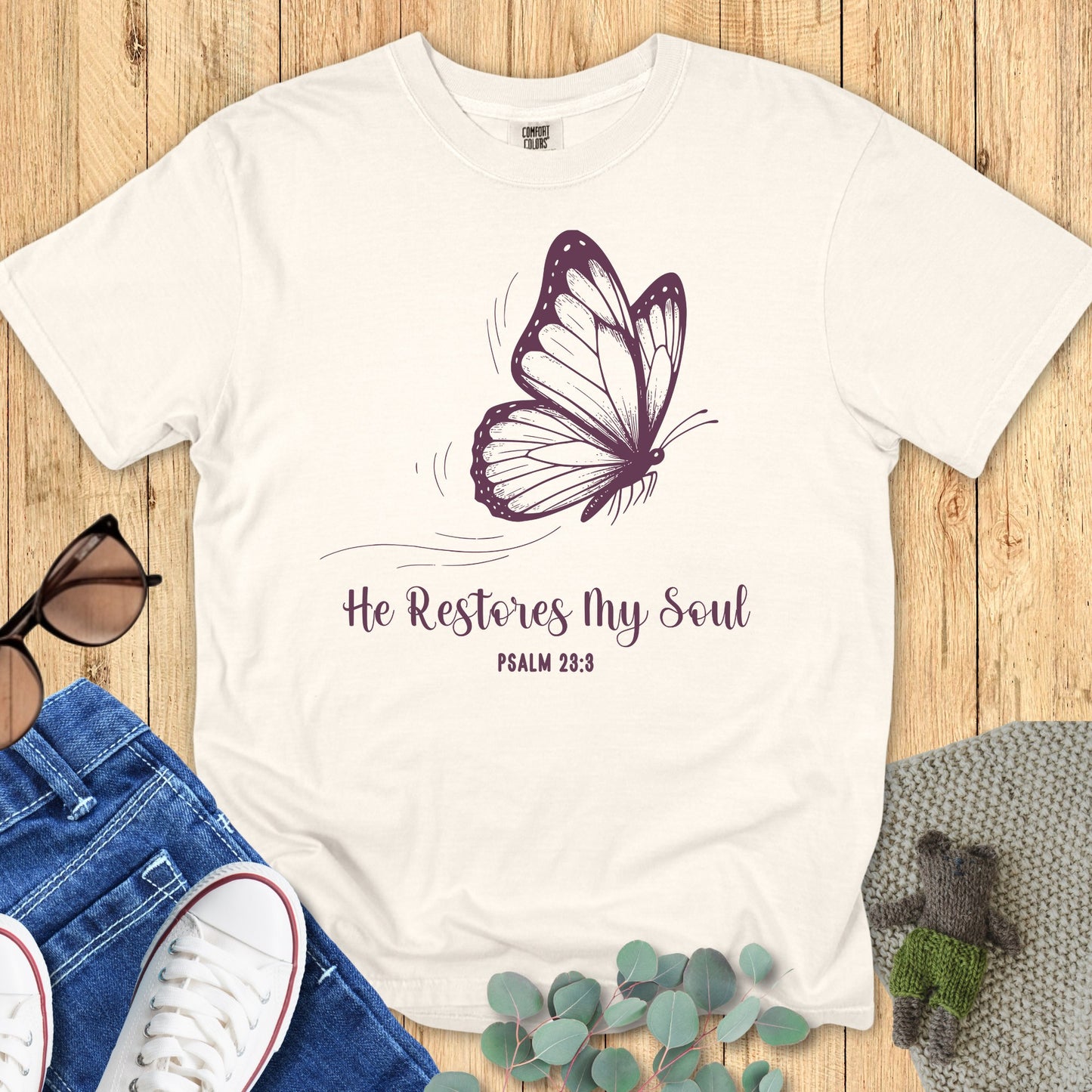 Flatlay of a Comfort Colors 1717 ivory “He Restores My Soul – Psalm 23:3” t-shirt featuring elegant purple butterfly design. A soft women’s scripture tee — uplifting faith-based apparel for women and modern Christian shirt, perfect for Christian mom gift ideas.