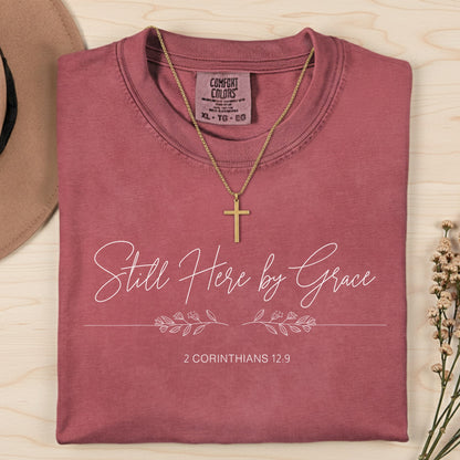 Flatlay of a Comfort Colors 1717 crimson “Still Here by Grace – 2 Corinthians 12:9” t-shirt with minimalist floral line design and delicate lettering. A graceful women’s scripture tee — uplifting faith-based apparel for women and modern Christian shirt, perfect Christian mom gift idea.