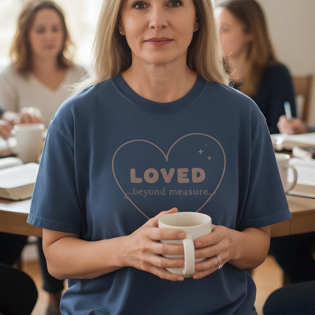 Woman holding a coffee mug in a Bible study group wearing a Comfort Colors 1717 midnight “Loved Beyond Measure – Ephesians 3:18-19” t-shirt. A gentle women’s scripture tee — heartfelt faith-based apparel for women and modern Christian shirt, perfect Christian mom gift idea.