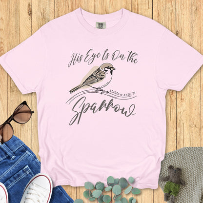 Flatlay of a Comfort Colors 1717 blossom “His Eye Is on the Sparrow – Matthew 10:29-31” t-shirt with minimal sparrow artwork and soft pastel tones. A feminine women’s scripture tee — gentle faith-based apparel for women and modern Christian shirt, sweet Christian mom gift idea.