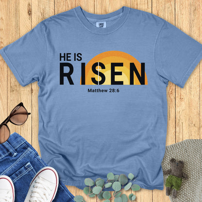 Flatlay of a Comfort Colors 1717 blue jean “He Is Risen – Matthew 28:6” t-shirt showcasing a warm yellow sunrise design. A classic women’s scripture tee — encouraging faith-based apparel for women and modern Christian shirt, inspiring Christian mom gift idea for Easter.