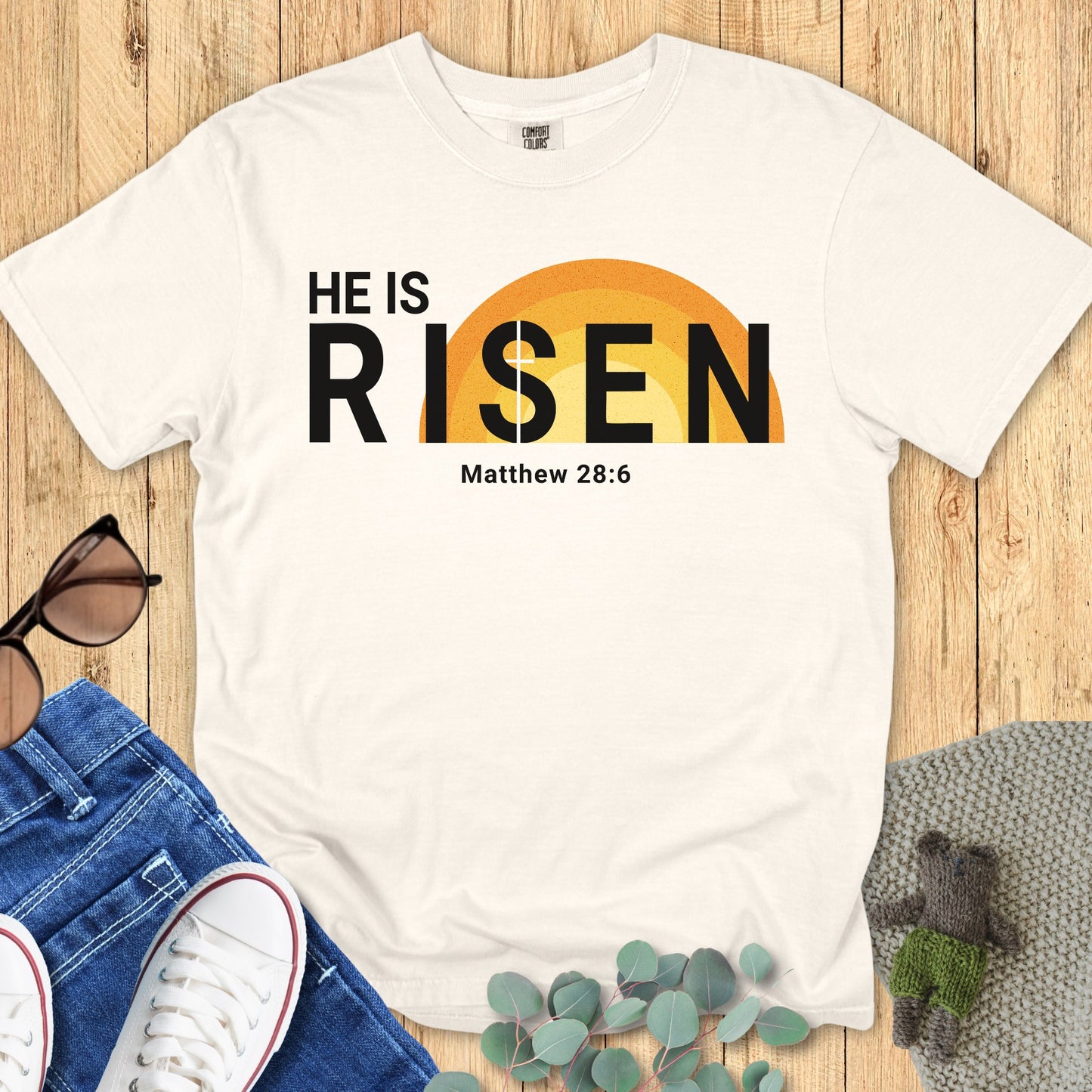 Flatlay of a Comfort Colors 1717 ivory “He Is Risen – Matthew 28:6” t-shirt featuring a bold sunrise graphic and minimalist typography. A bright women’s scripture tee — faith-based apparel for women and modern Christian shirt, thoughtful Christian mom gift idea for Easter.