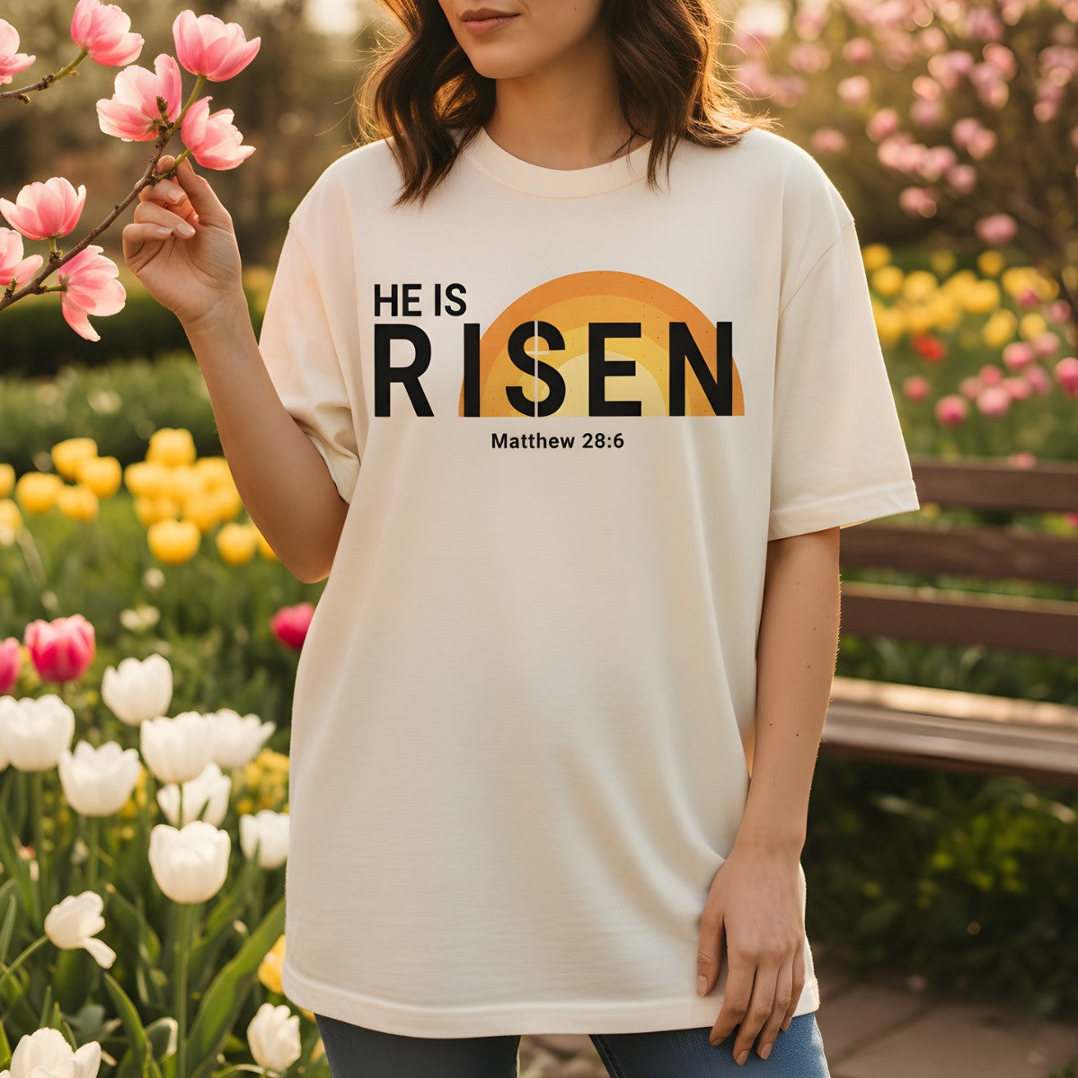 Woman standing in a garden of tulips wearing a Comfort Colors 1717 ivory “He Is Risen – Matthew 28:6” t-shirt with a sunrise design. A joyful women’s scripture tee — uplifting faith-based apparel for women and modern Christian shirt, perfect Easter or Christian mom gift idea.