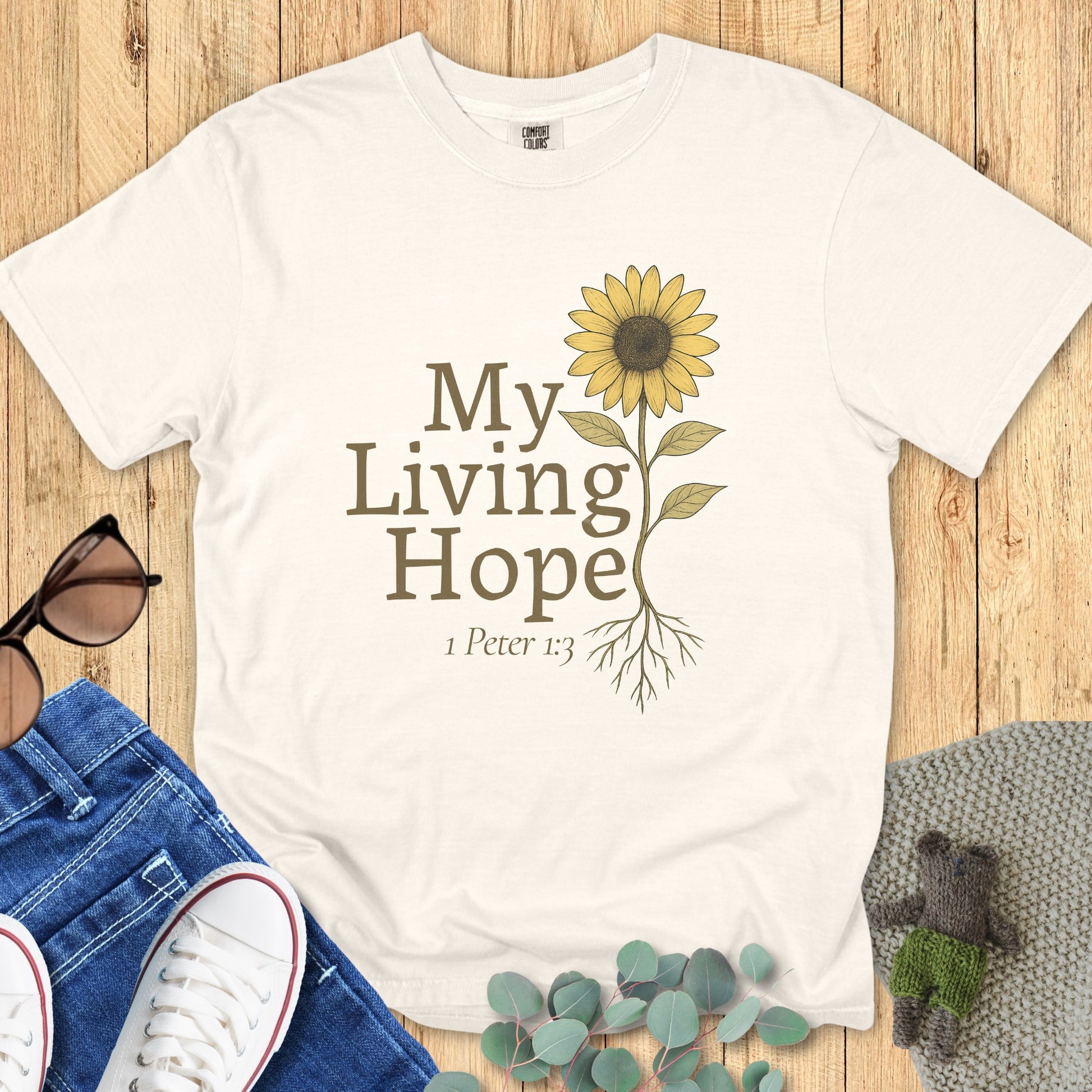 Flatlay of a Comfort Colors 1717 ivory “My Living Hope – 1 Peter 1:3” t-shirt with hand-drawn sunflower and rooted design. A beautiful women’s scripture tee — faith-based apparel for women and modern Christian shirt, perfect Christian mom gift idea.