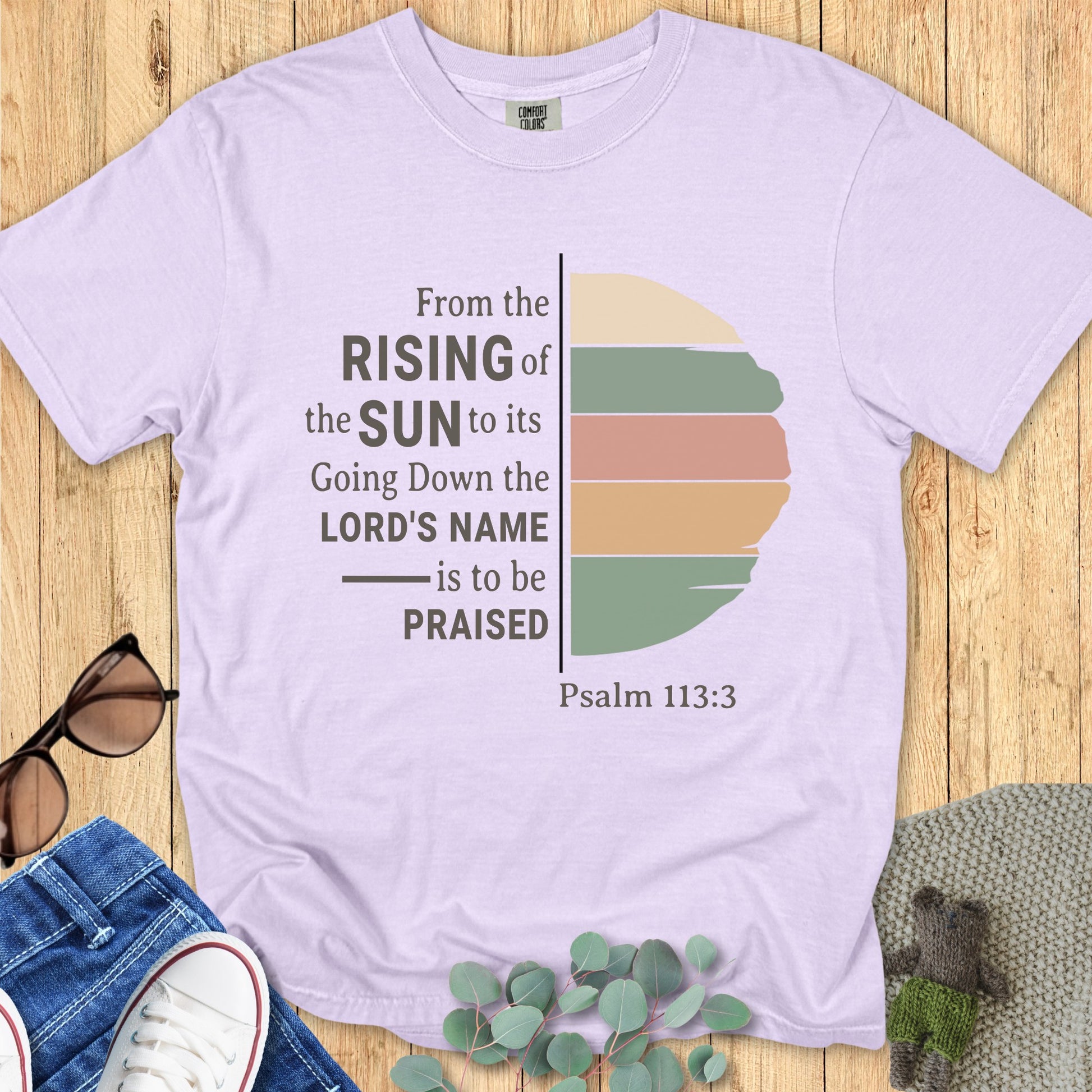 Flatlay of a Comfort Colors 1717 orchid “From the Rising of the Sun to its Going Down, the Lord’s Name is to be Praised – Psalm 113:3” t-shirt with muted sunrise graphic. A soft and inspiring women’s scripture tee — faith-based apparel for women and modern Christian shirt, lovely Christian mom gift idea.
