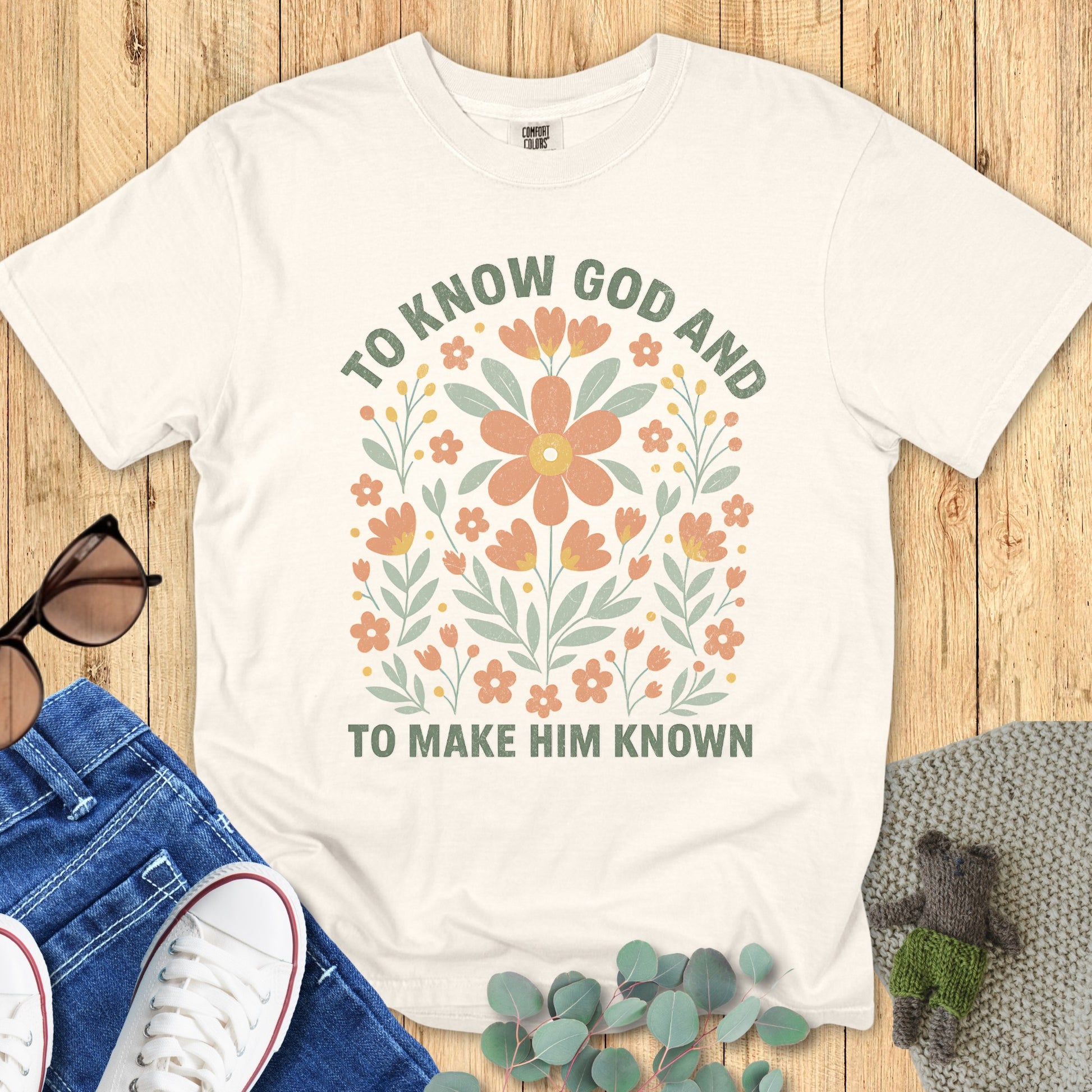 Flatlay of a Comfort Colors 1717 ivory “To Know God and To Make Him Known” t-shirt with soft pastel floral design. A beautiful women’s scripture tee — faith-based apparel for women and modern Christian shirt, ideal Christian mom gift idea.