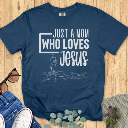 Flatlay of a Comfort Colors 1717 midnight “Just a Mom Who Loves Jesus” t-shirt featuring a serene outline of Jesus walking on the water. A cozy women’s scripture tee — faith-based apparel for women and modern Christian shirt, lovely Christian mom gift idea.