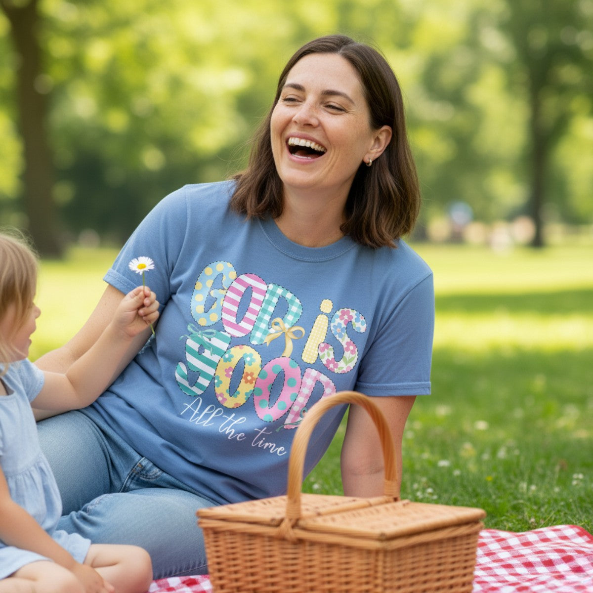 Mom laughing with her child during a sunny picnic while wearing a Comfort Colors 1717 blue “God Is Good All the Time” t-shirt with colorful patchwork letters and bows. A joyful women’s scripture tee — faith-based apparel for women and modern Christian shirt, thoughtful Christian mom gift idea.