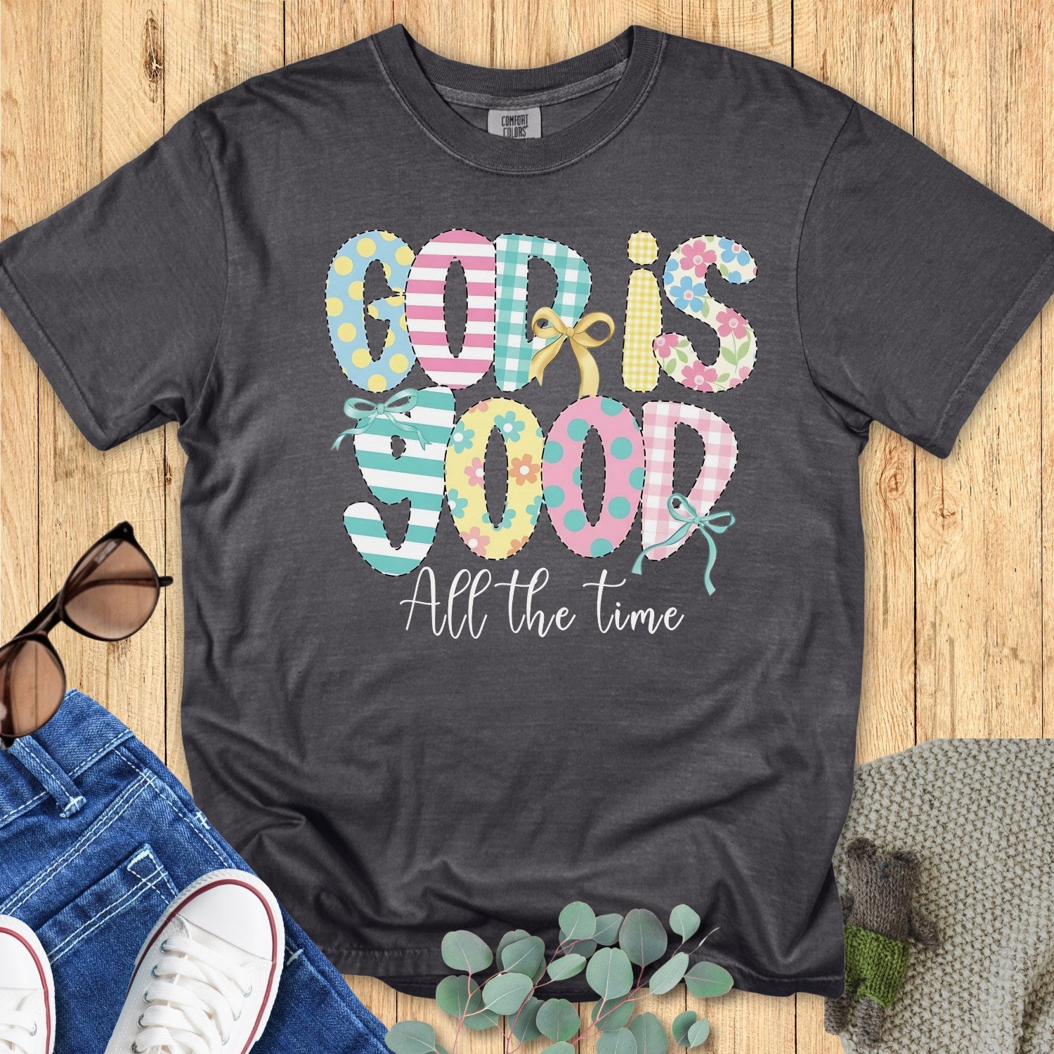 Flatlay of a Comfort Colors 1717 pepper “God Is Good All the Time” t-shirt with colorful stitched-style lettering and cute bows. A soft and inspiring women’s scripture tee — faith-based apparel for women and modern Christian shirt, meaningful Christian mom gift idea.