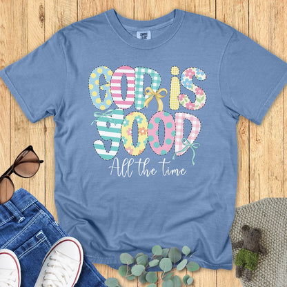 Flatlay of a Comfort Colors 1717 blue jean “God Is Good All the Time” t-shirt featuring pastel patchwork lettering with playful bow accents. A bright and joyful women’s scripture tee — faith-based apparel for women and modern Christian shirt, ideal Christian mom gift idea.