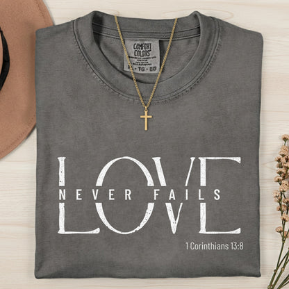 Folded Comfort Colors 1717 pepper “Love Never Fails” t-shirt styled with a gold cross necklace and soft neutral tones. A classic women’s scripture tee — faith-based apparel for women and modern Christian shirt referencing 1 Corinthians 13:8, elegant Christian mom gift idea.