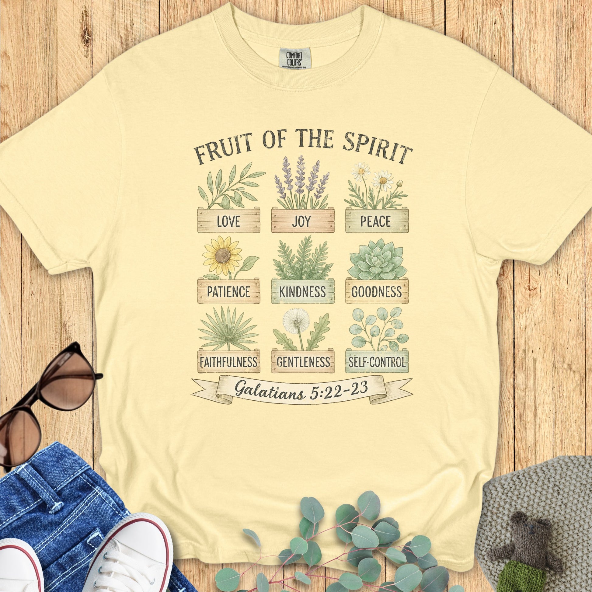 Flatlay of a Comfort Colors 1717 butter “Fruit of the Spirit” t-shirt featuring gentle botanical artwork and scripture from Galatians 5:22–23. A light and uplifting women’s scripture tee — faith-based apparel for women and modern Christian shirt, thoughtful Christian mom gift idea.