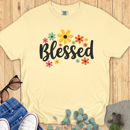 Flatlay of a Comfort Colors 1717 butter “Blessed” t-shirt with colorful floral graphics surrounding the word “Blessed.” A happy women’s scripture tee — faith-based apparel for women and modern Christian shirt, ideal Christian mom gift idea.