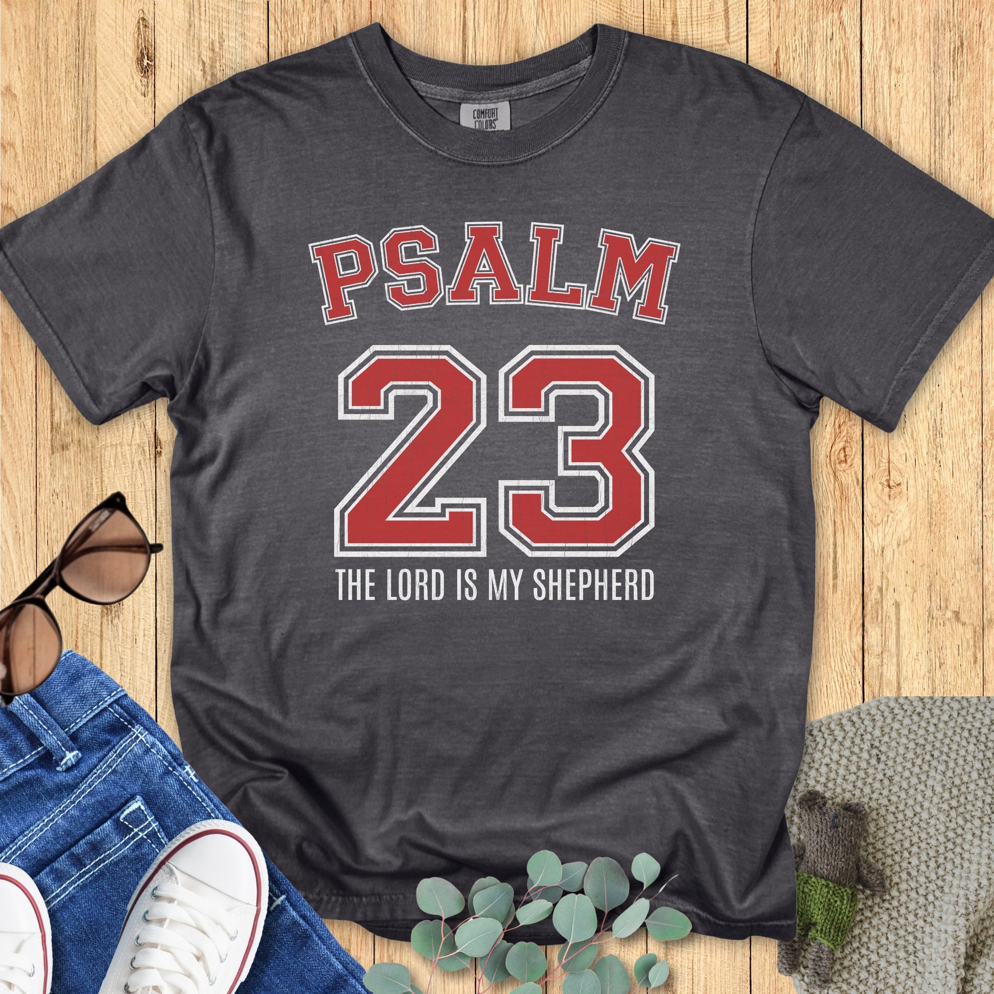 Flatlay of a Comfort Colors 1717 pepper gray “Psalm 23 The Lord is My Shepherd” t-shirt with red varsity numbers and collegiate style design. A casual women’s scripture tee — faith-based apparel for women and modern Christian shirt, meaningful Christian mom gift idea.