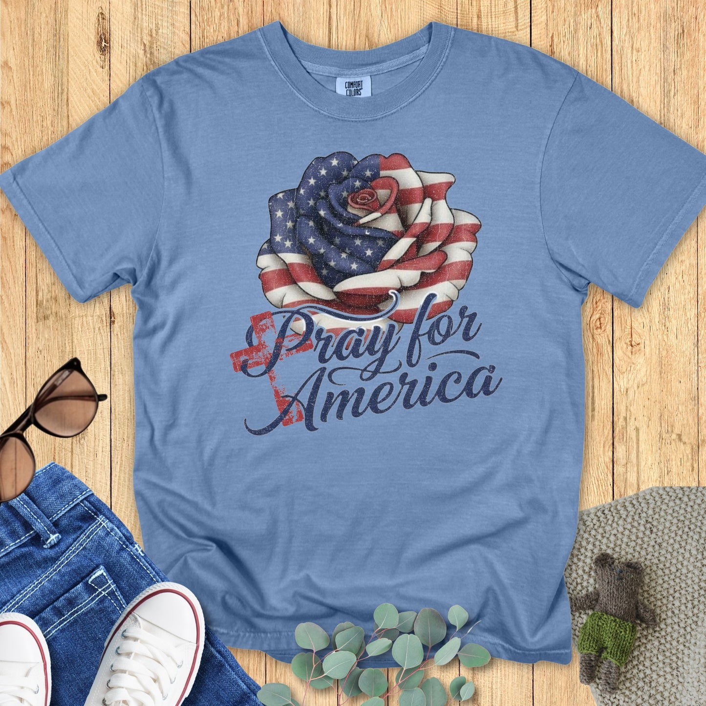 Flatlay of a Comfort Colors 1717 blue jean “Pray for America” t-shirt with vintage-style red, white, and blue rose graphic. A soft women’s scripture tee — faith-based apparel for women and modern Christian shirt, thoughtful Christian mom gift idea for patriotic holidays.