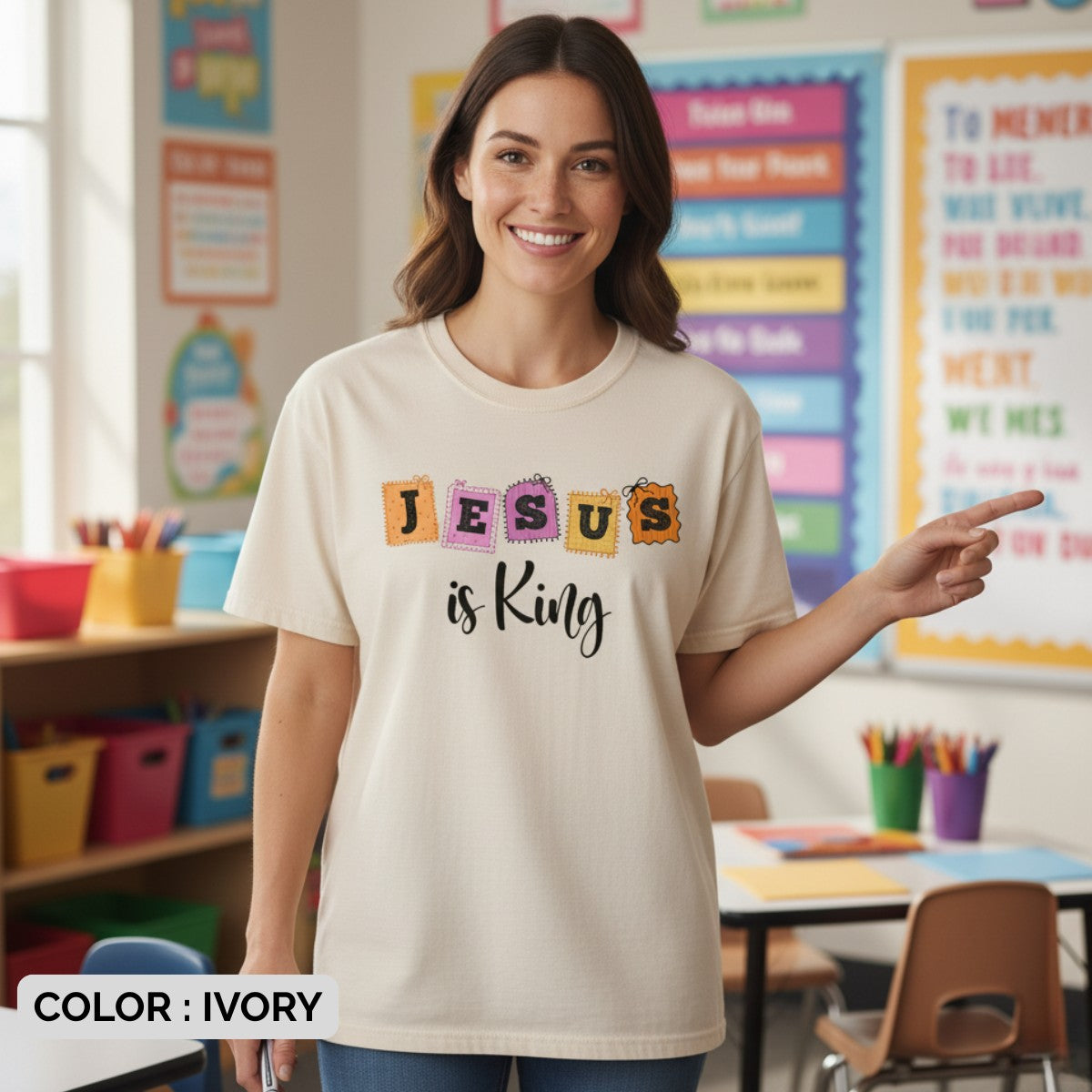 Smiling woman wearing a Comfort Colors 1717 ivory “Jesus is King” t-shirt with colorful patch-style lettering, standing in a cheerful classroom. A bright women’s scripture tee - faith-based apparel for women and modern Christian shirt, thoughtful Christian mom gift idea for teachers and believers alike.