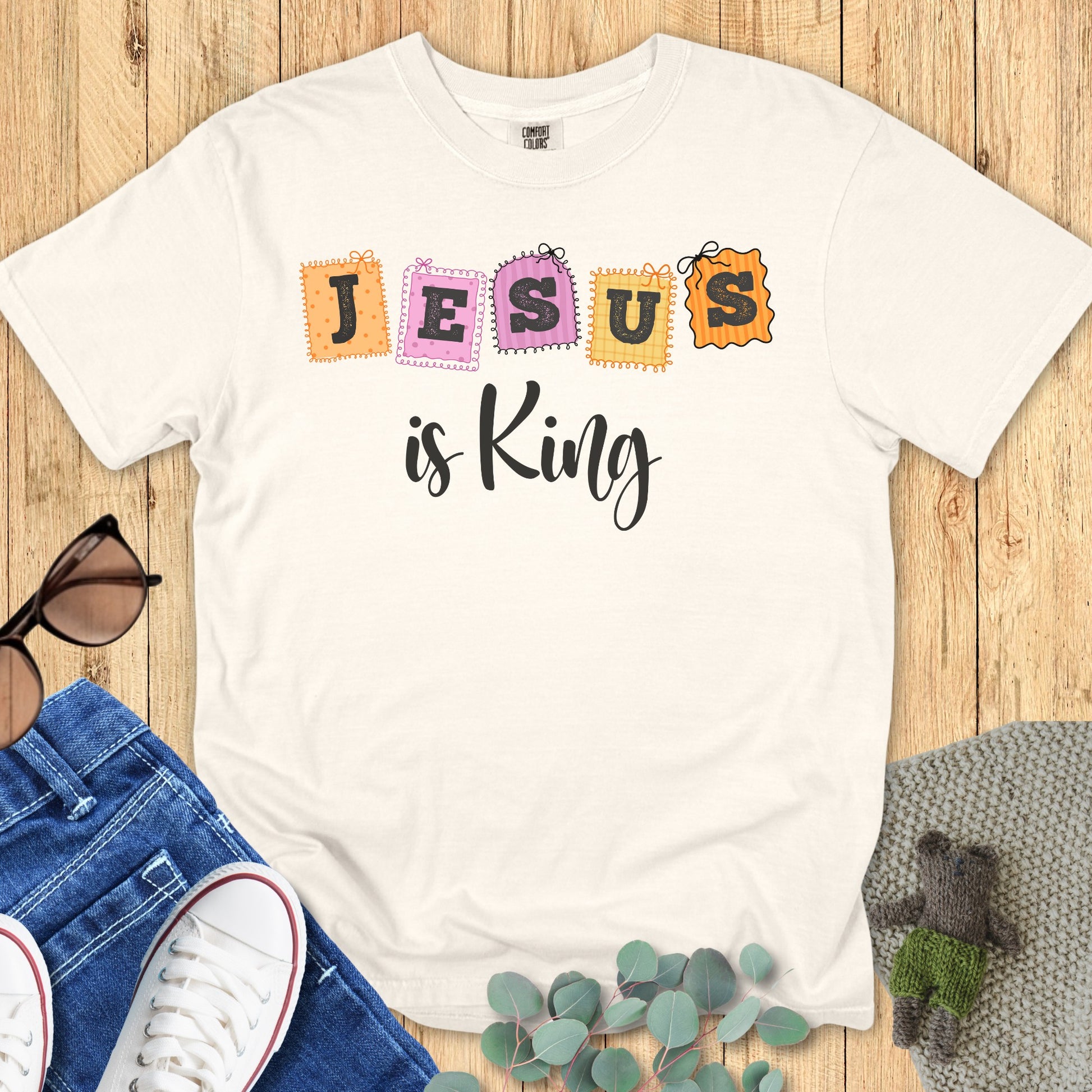Flatlay of a Comfort Colors 1717 ivory “Jesus is King” t-shirt featuring pastel stitched block letters. A classic women’s scripture tee - faith-based apparel for women and modern Christian shirt, beautiful Christian mom gift idea for daily inspiration.