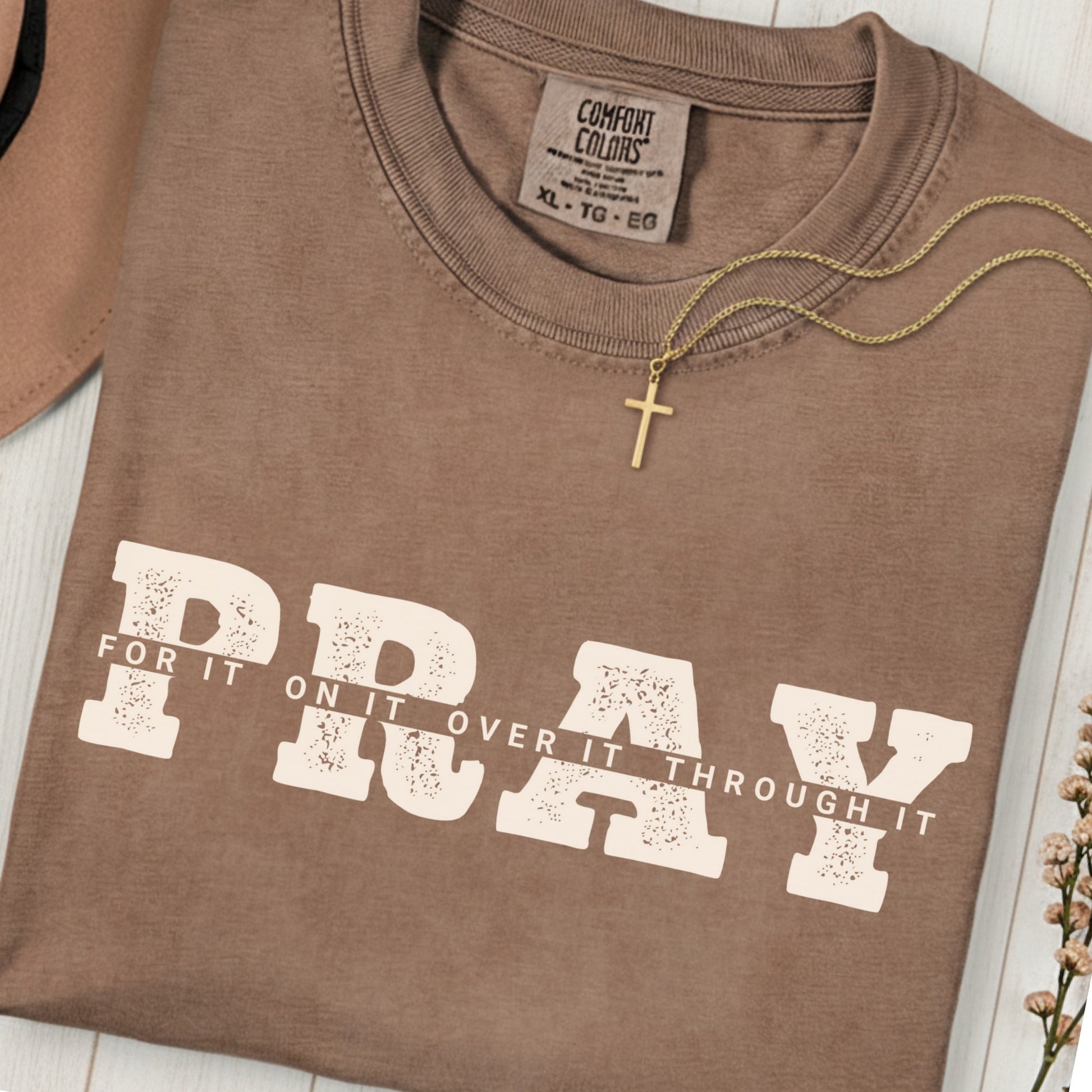 Folded Comfort Colors 1717 espresso “PRAY for it, on it, over it, through it” t-shirt with gold cross necklace on top. A classic women’s scripture tee -  faith-based apparel for women and modern Christian shirt, ideal Christian mom gift idea and prayer reminder.
