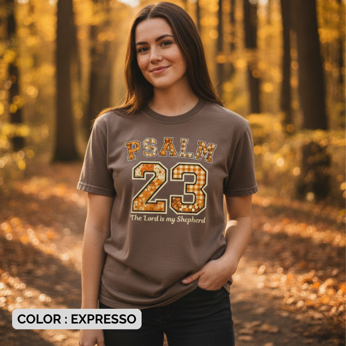 Woman wearing a Comfort Colors 1717 expresso “Psalm 23 — The Lord is my Shepherd” t-shirt with retro floral and gingham patch details, standing in a fall forest. A warm women’s scripture tee - faith-based apparel for women and modern Christian shirt, thoughtful Christian mom gift idea.