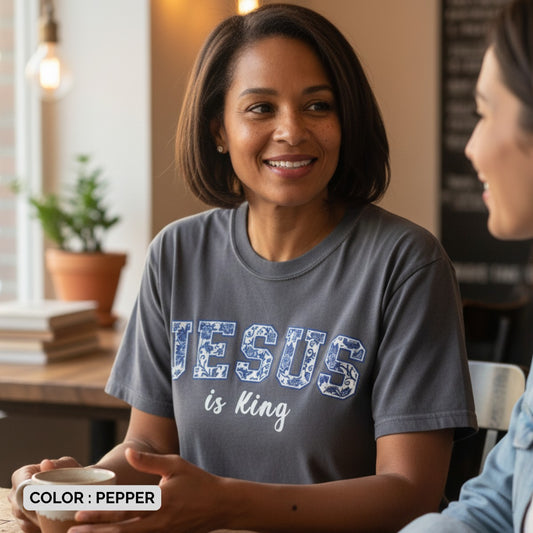 Woman wearing a Comfort Colors 1717 pepper “Jesus is King” t-shirt with blue floral varsity text while enjoying coffee indoors. A relaxed women’s scripture tee - faith-based apparel for women and modern Christian shirt, uplifting Christian mom gift idea for daily inspiration.