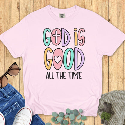 Flatlay of a Comfort Colors 1717 blossom pink “God Is Good All The Time” t-shirt in cute pastel lettering with cross and heart icons. A sweet women’s scripture tee -  faith-based apparel for women and modern Christian shirt, ideal Christian mom gift idea for daily faith wear.