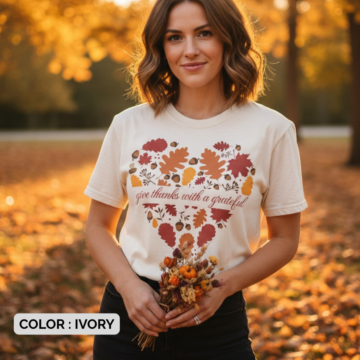 Woman wearing a Comfort Colors 1717 ivory “Give Thanks with a Grateful Heart” t-shirt with fall leaf heart design, standing in a golden autumn park. A soft women’s scripture tee - faith-based apparel for women and modern Christian shirt, heartfelt Christian mom gift idea for Thanksgiving season.