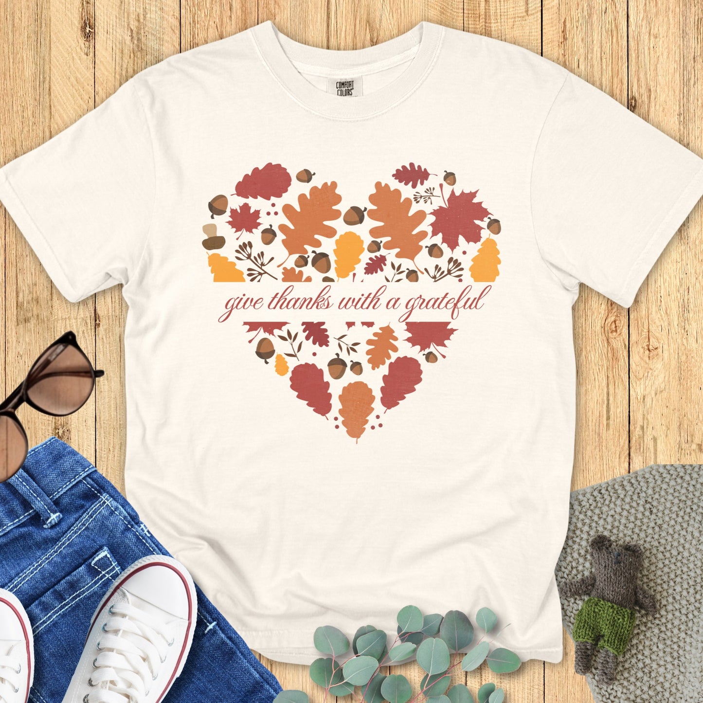 Flatlay of a Comfort Colors 1717 ivory “Give Thanks with a Grateful Heart” t-shirt decorated with autumn leaves and acorns forming a heart shape. A minimalist women’s scripture tee - faith-based apparel for women and modern Christian shirt, lovely Christian mom gift idea for everyday gratitude.