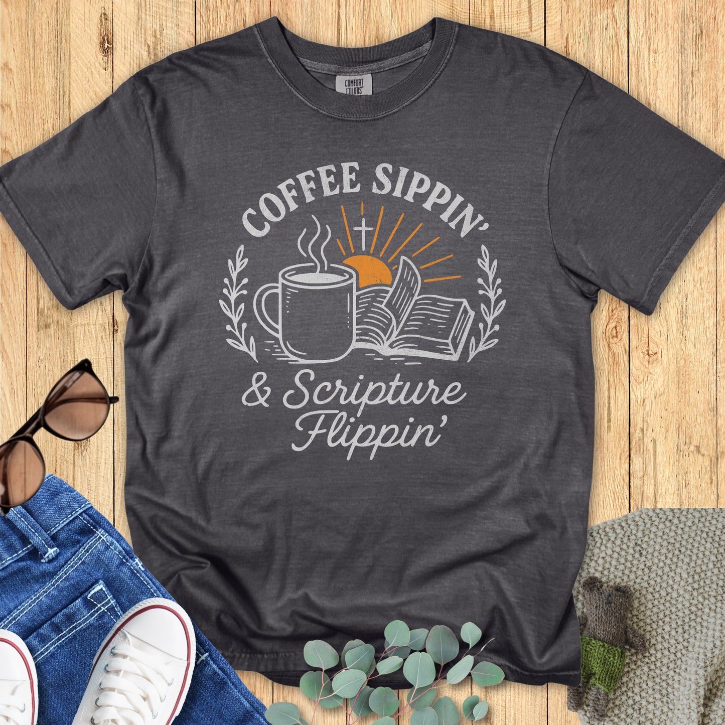 Flatlay of a Comfort Colors 1717 pepper “Coffee Sippin’ & Scripture Flippin’” t-shirt featuring a steaming mug, open Bible, and sunrise graphic. A stylish women’s scripture tee - faith-based apparel for women and modern Christian shirt, thoughtful Christian mom gift idea for coffee and Bible lovers.
