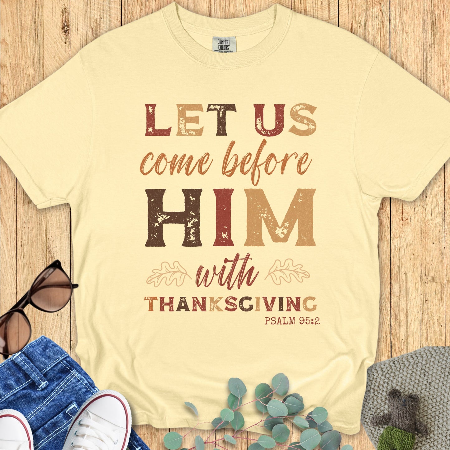 Butter-colored Christian shirt flat lay featuring the verse “Let Us Come Before Him With Thanksgiving, Psalm 95:2.” Warm fall tones, distressed text, autumn leaf elements, Christian Thanksgiving apparel for women.