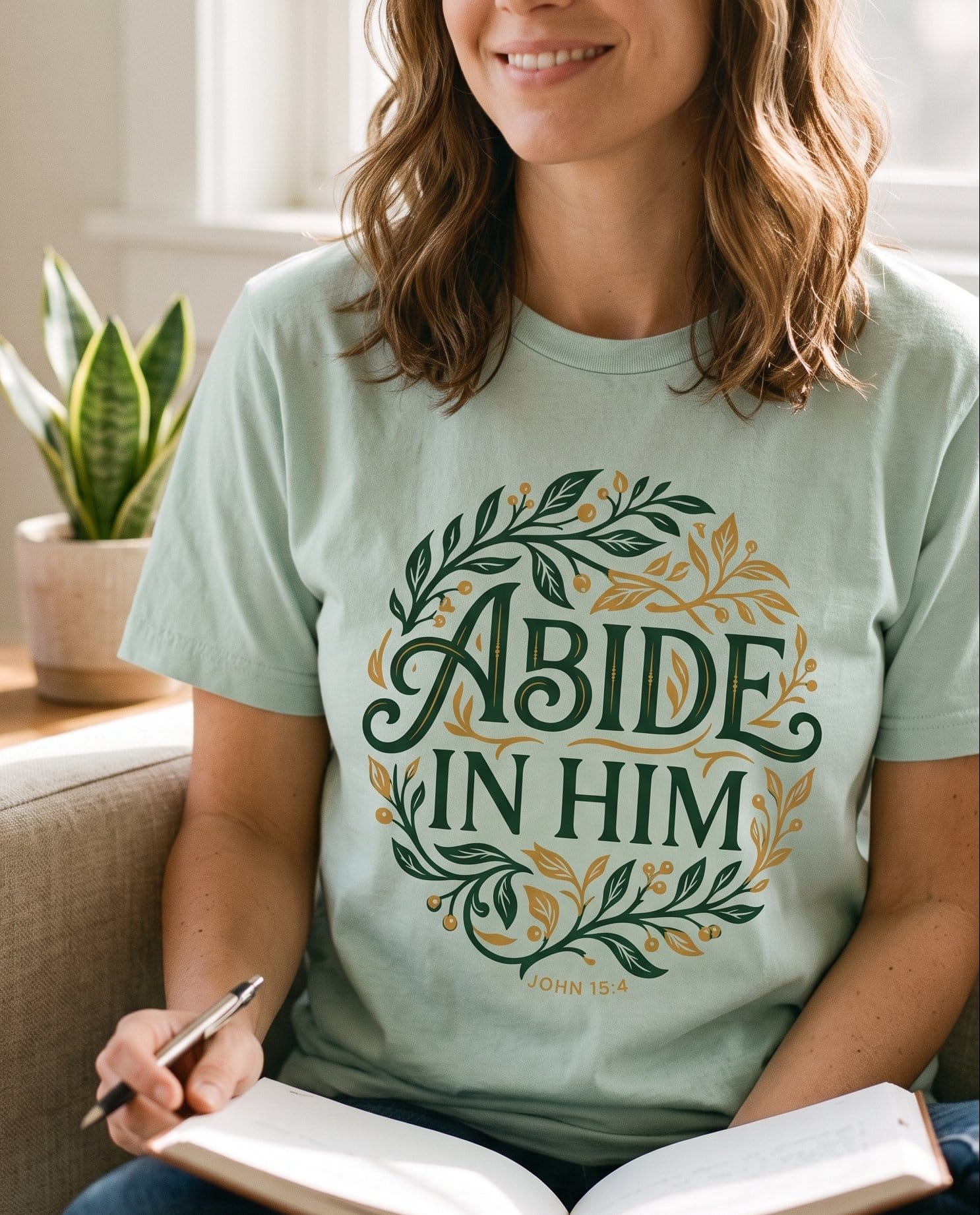 Abide in Him John 15:4 – Bay women's Christian tee, lifestyle photo in cozy living room – Deep green and warm gold botanical wreath encircling ornate 'Abide in  – John 15:4 – She Wears Faith