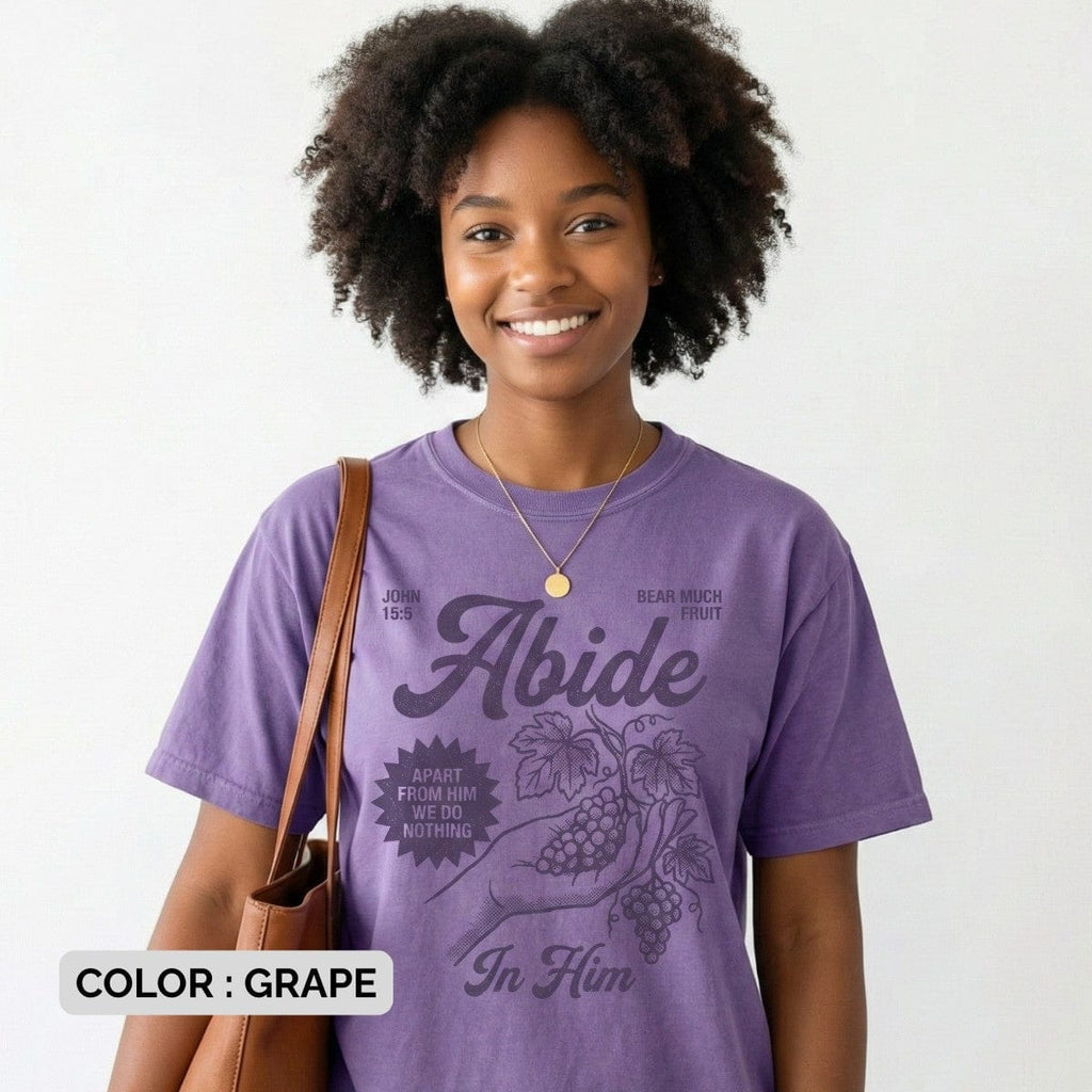 Smiling woman wearing a Grape Purple Comfort Colors 1717 tshirt featuring a vintage illustration of a hand holding grapes. The design reads "Abide In Him" with John 15:5 scripture, making it a meaningful Christian women tee for US faith wear shops.