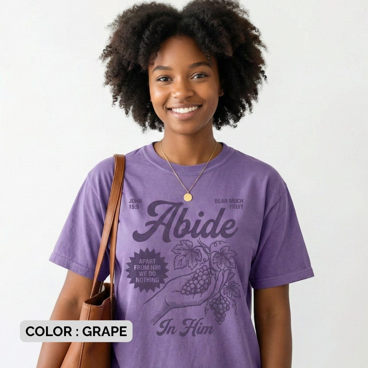Smiling woman wearing a Grape Purple Comfort Colors 1717 tshirt featuring a vintage illustration of a hand holding grapes. The design reads "Abide In Him" with John 15:5 scripture, making it a meaningful Christian women tee for US faith wear shops.