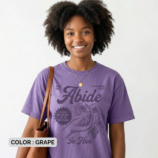 Smiling woman wearing a Grape Purple Comfort Colors 1717 tshirt featuring a vintage illustration of a hand holding grapes. The design reads "Abide In Him" with John 15:5 scripture, making it a meaningful Christian women tee for US faith wear shops.