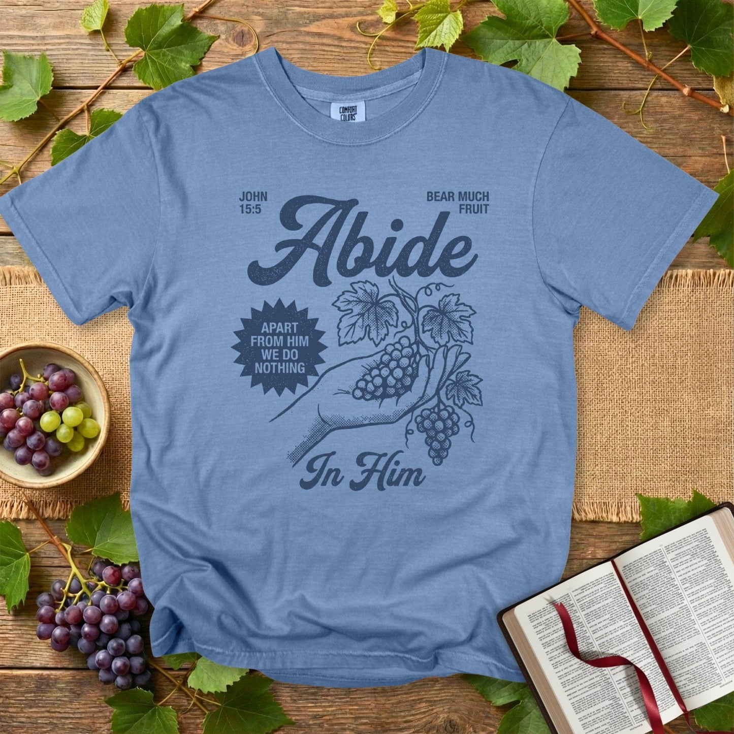 Comfort Colors 1717 blue jean Christian women tshirt with the “Abide in Him” John 15:5 grape vine artwork, vintage hand-drawn style. Faith-based US faith wear tee shown in a flat-lay setup surrounded by fresh grapes and an open Bible.