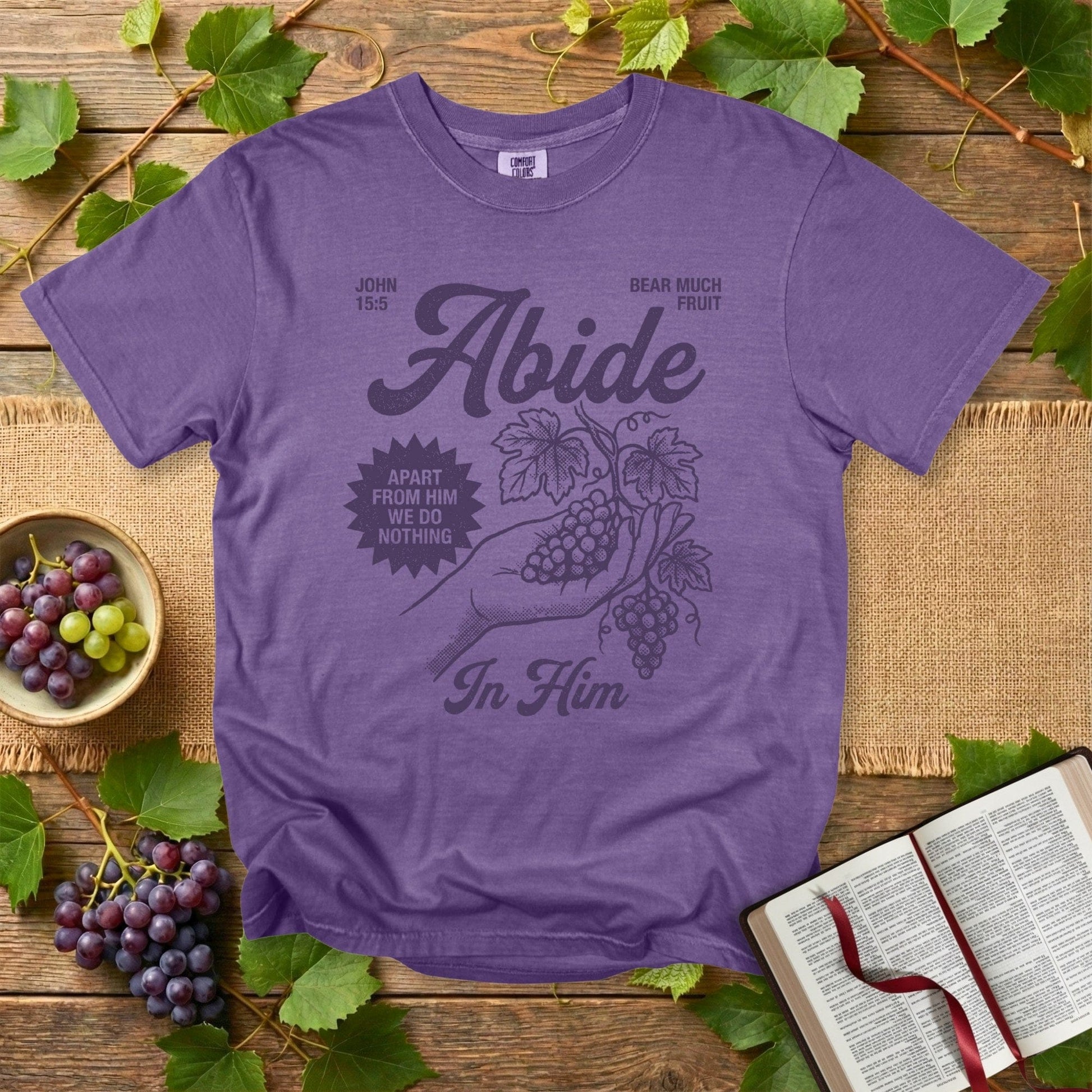 Comfort Colors 1717 grape purple Christian women tee featuring the “Abide in Him” John 15:5 scripture design with grapes and hand illustration. Stylish Christian tshirt for US faith wear, displayed in a flat-lay with grapes, leaves, and an open Bible.