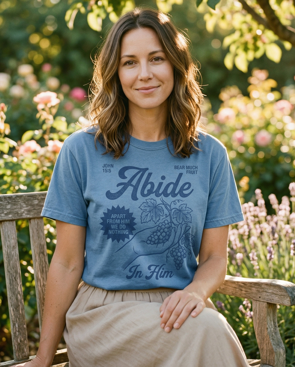 A woman sitting on a wooden bench in a flowering garden, wearing a blue short-sleeved t-shirt that displays the text "John 15:5," "Abide," and "Apart from Him we do nothing" with an illustration of grapes