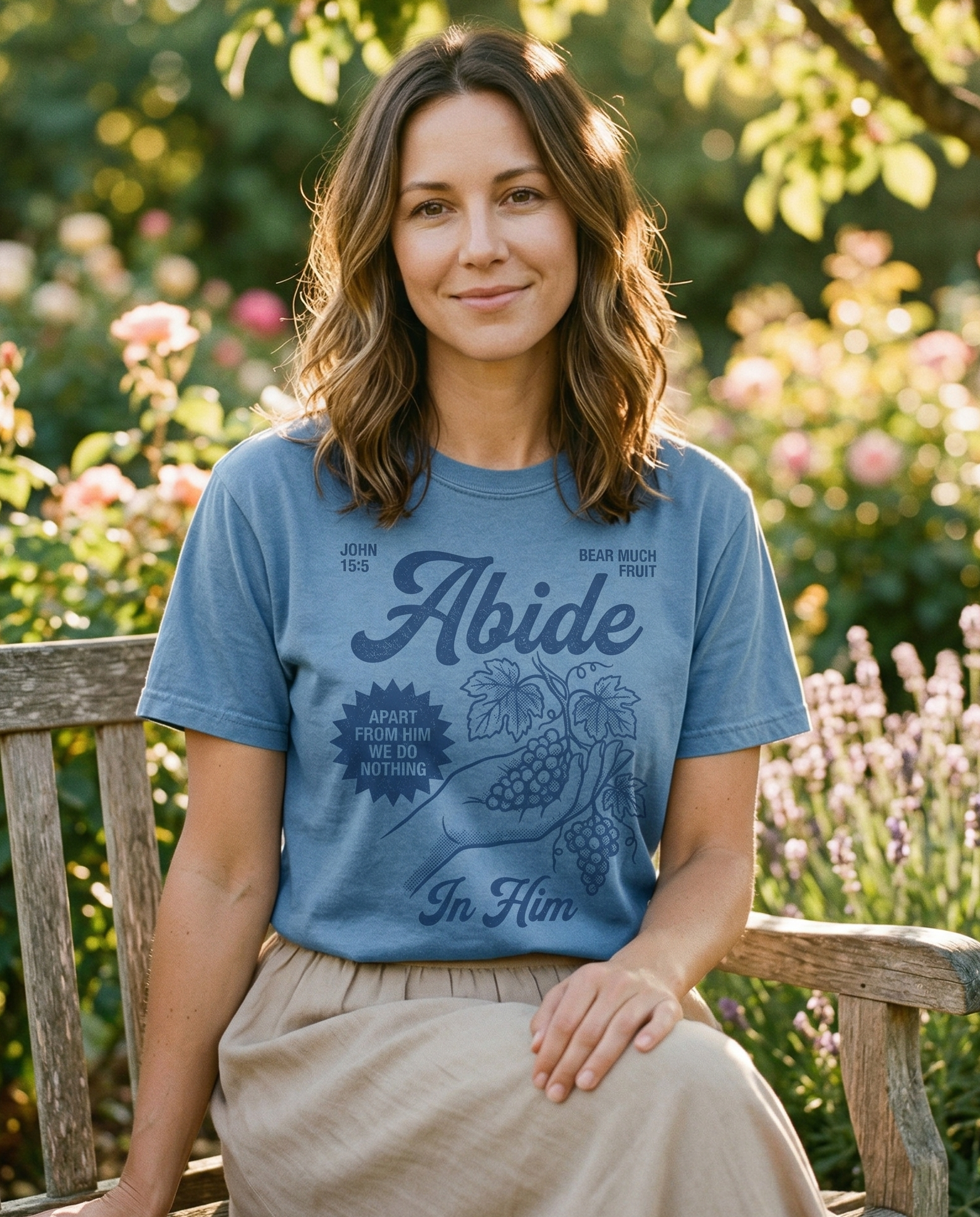 A woman sitting on a wooden bench in a flowering garden, wearing a blue short-sleeved t-shirt that displays the text "John 15:5," "Abide," and "Apart from Him we do nothing" with an illustration of grapes