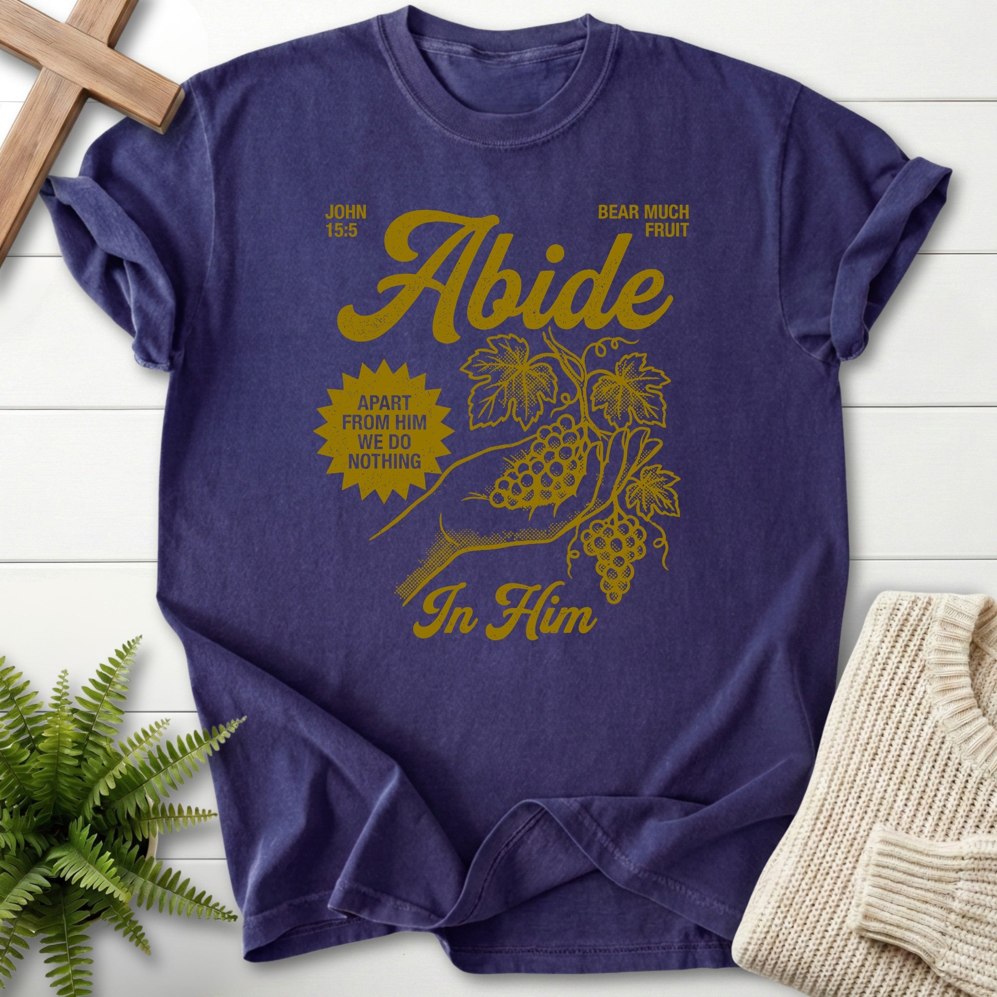 Purple t-shirt with yellow graphic and text on a white wooden surface with a plant and sweater.