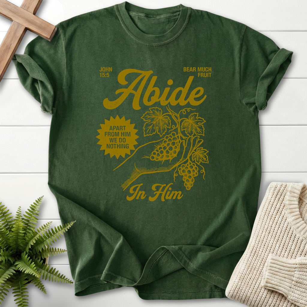 Green t-shirt with yellow text and graphics on a white wooden surface with a plant and sweater.