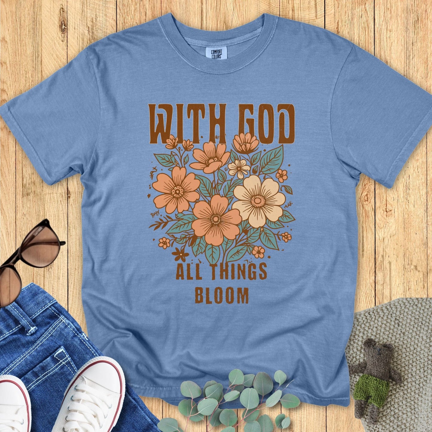 hristian women’s Comfort Colors 1717 t-shirt in Blue Jean with floral design and the words ‘With God All Things Bloom.’ Displayed flat lay with jeans, sneakers, and sunglasses on a wood background.