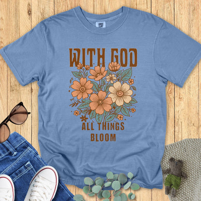 hristian women’s Comfort Colors 1717 t-shirt in Blue Jean with floral design and the words ‘With God All Things Bloom.’ Displayed flat lay with jeans, sneakers, and sunglasses on a wood background.