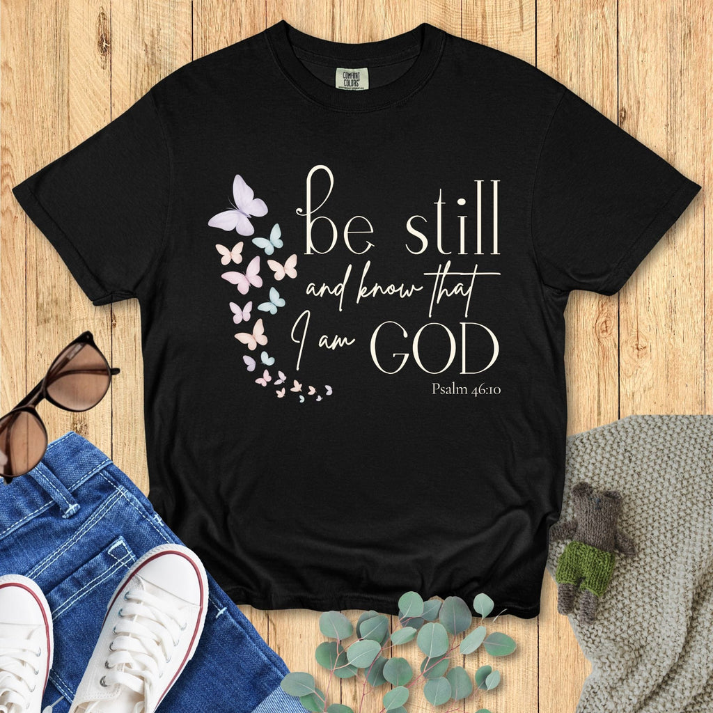 Black Comfort Colors Christian T-shirt with butterfly design and scripture "Be Still and Know That I Am God, Psalm 46:10." Faith-based inspirational tee flat lay with jeans, sneakers, and sunglasses on wood background.