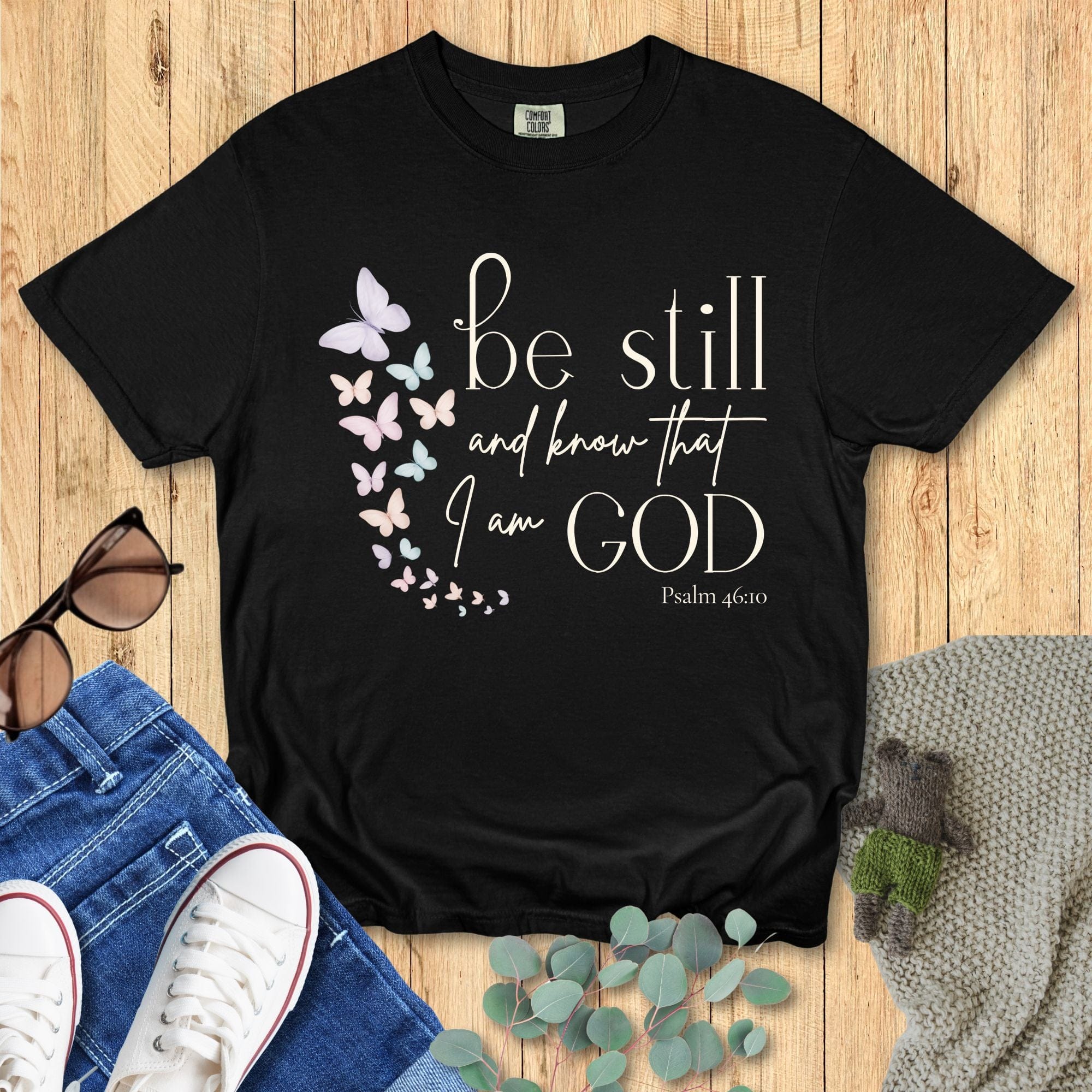 Black Comfort Colors Christian T-shirt with butterfly design and scripture "Be Still and Know That I Am God, Psalm 46:10." Faith-based inspirational tee flat lay with jeans, sneakers, and sunglasses on wood background.
