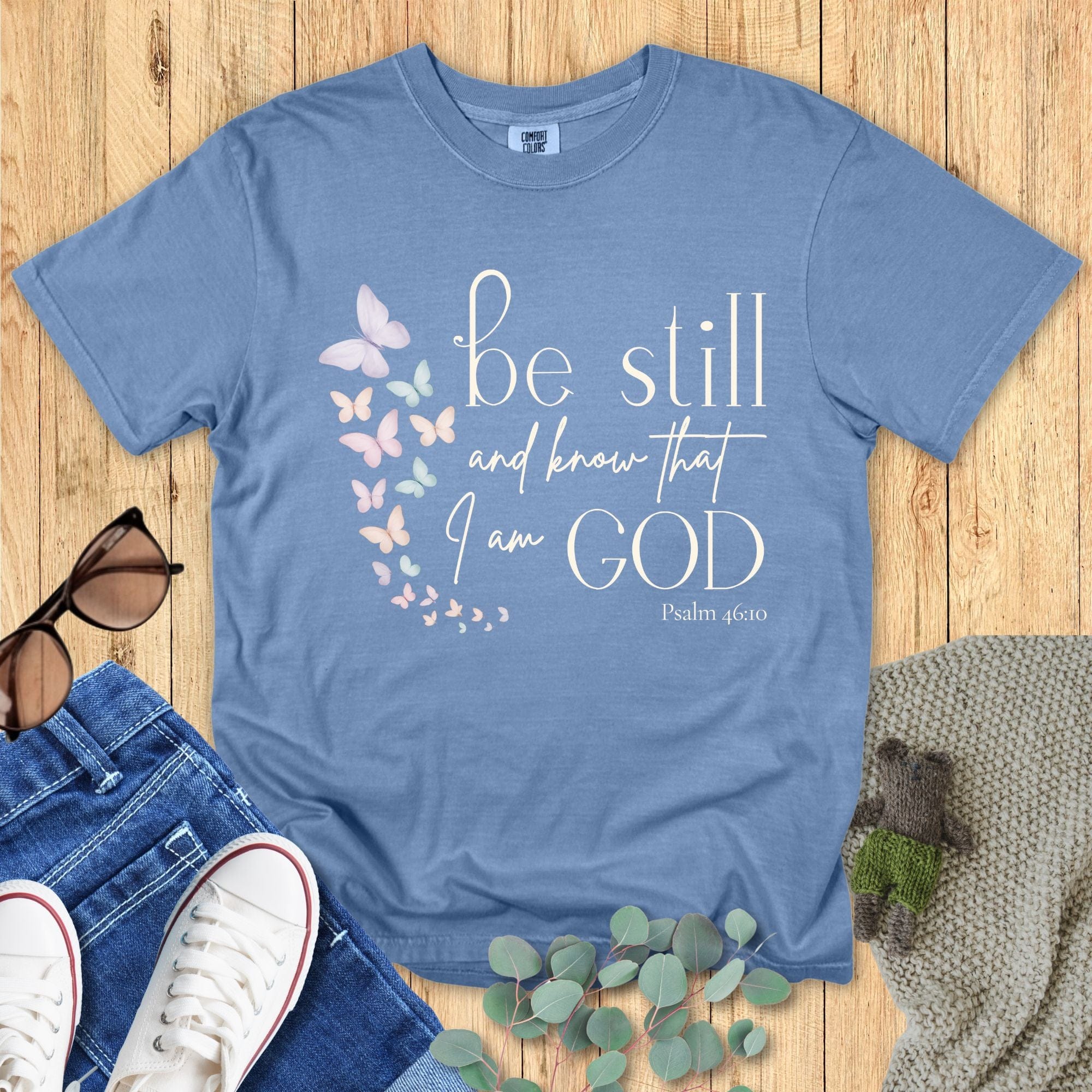 Blue Jean Comfort Colors Christian T-shirt featuring pastel butterfly artwork and verse "Be Still and Know That I Am God, Psalm 46:10." Religious graphic tee flat lay with casual outfit accessories on wooden surface.