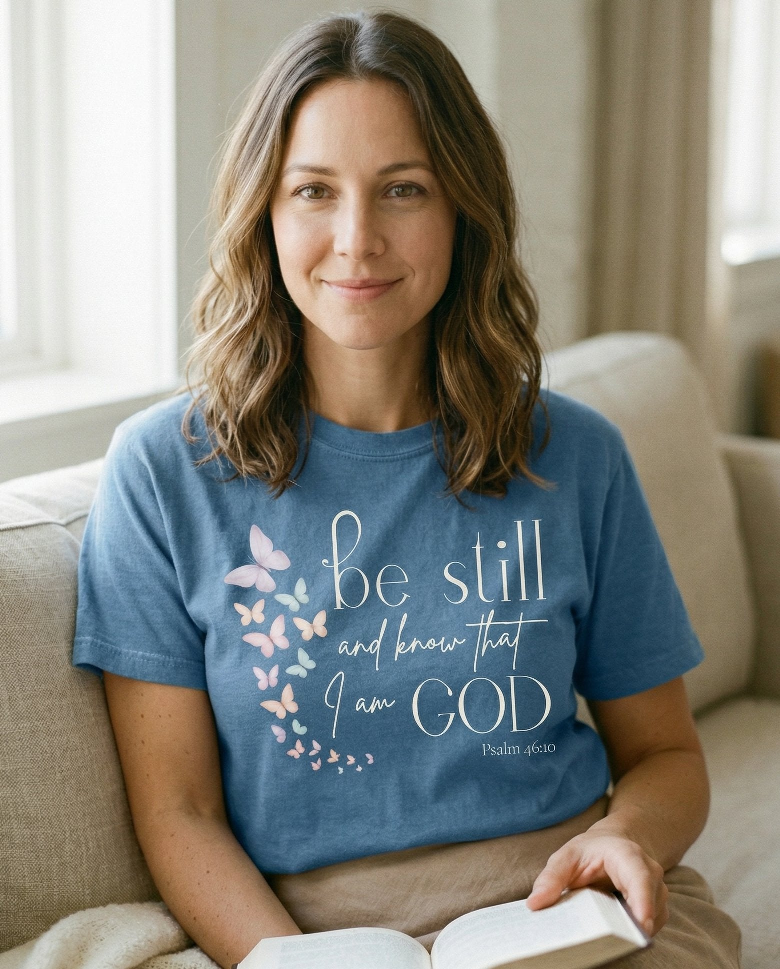 Be Still and Know Butterflies – Blue Jean women's Christian tee, lifestyle photo in cozy living room – Soft watercolor butterflies in pastel pink – Psalm 46:10 – She Wears Faith