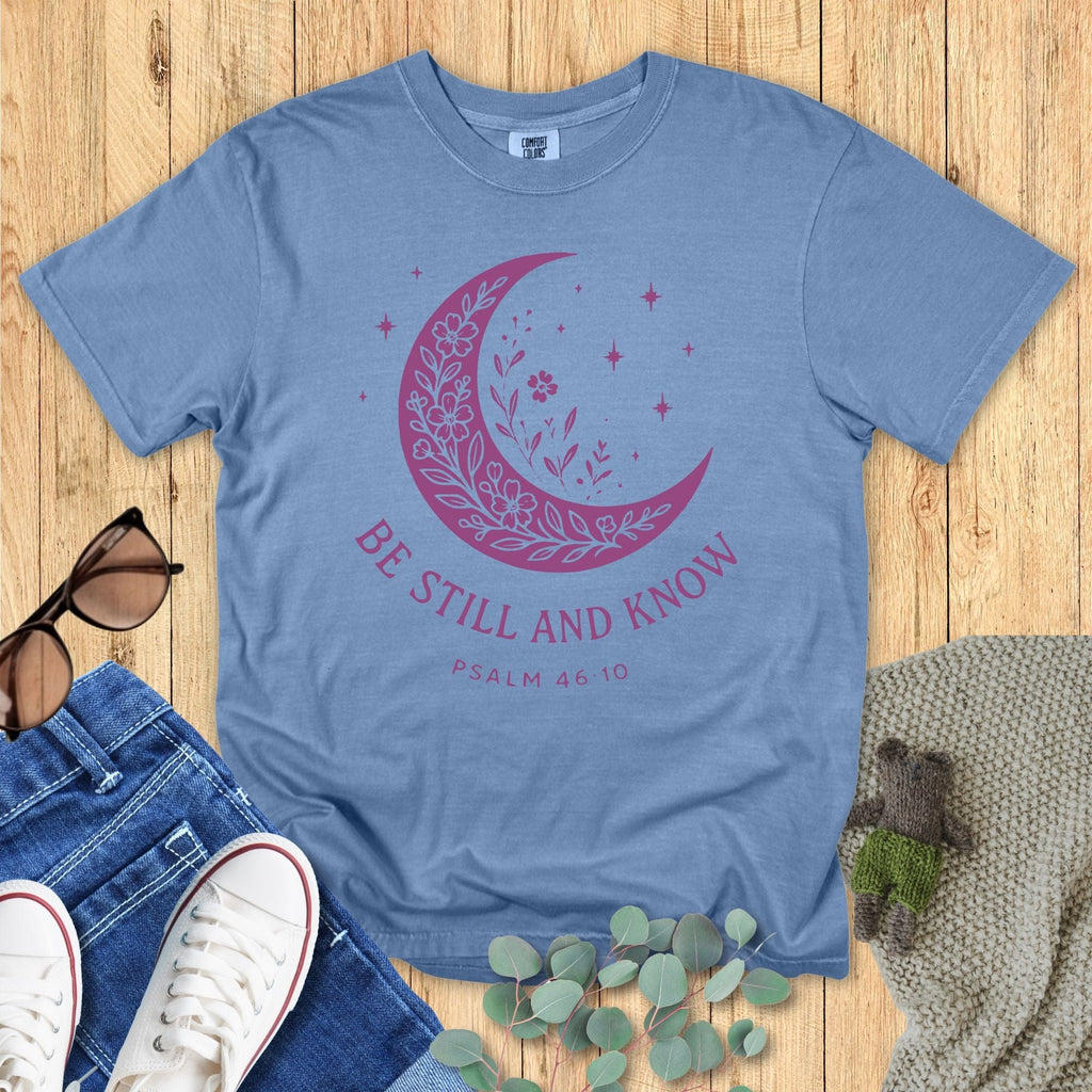 Flat lay of a Comfort Colors Christian T-shirt in blue jean, featuring a purple crescent moon filled with floral details and stars, with text “Be Still and Know – Psalm 46:10.”